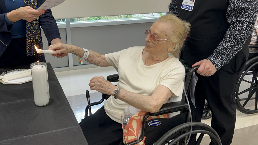 An elderly woman in a wheelchair lighting a white candle on a table, assisted by a man standing beside her. Another person stands by with a paper in hand.